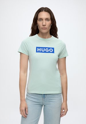 Femme aux longs cheveux bruns ondulés portant un t-shirt vert clair à manches courtes avec le logo bleu "HUGO" et un jean bleu clair, debout devant un fond blanc.