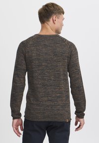Strikkegenser i mørkebrun og svart, med et teksturert mønster, lange ermer og rund hals. Merkelig accenttag nederst.
