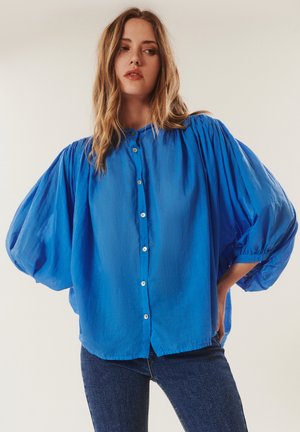 Femme portant une blouse ample bleu vif boutonnée avec des manches volumineuses et un jean bleu foncé, debout avec une main sur la hanche.