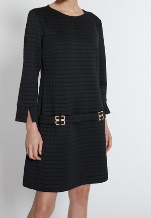 Robe noire texturée à manches longues avec motif carré, encolure ronde, fentes aux poignets et deux boucles décoratives dorées près des hanches.
