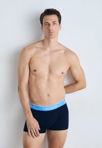 Boxers pour hommes en bleu marine avec une ceinture élastique bleu clair, portant le logo "Lacoste". Tissu lisse et design ajusté.