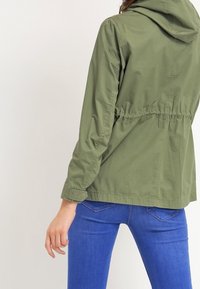 Olivgrön lättviktsjacka med huva, elastisk midja och lång ärm, med en slät textur och avslappnad design.