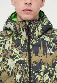 Vattert jakke med et grønt, svart og beige kamuflasjemønster. Den har glidelås, hette og en logo på brystet. Glatt tekstur.