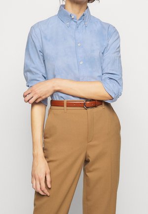 Osoba w jasno niebieskiej koszuli na guziki z podwiniętymi rękawami, beżowych spodniach z wysokim stanem i brązowym skórzanym pasku, z założonymi na piersi rękami.