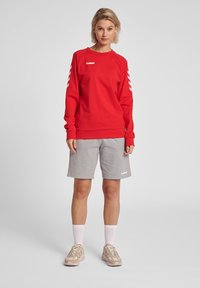 Rode sweatshirt met een wit logo en chevron-detail, gedragen met grijze shorts en beige sneakers, voorzien van witte accenten en geribbeld manchet.