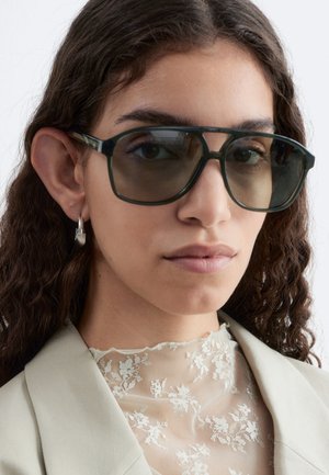 Jeune femme aux cheveux foncés ondulés portant de grandes lunettes de soleil foncées, des boucles d'oreilles argentées en anneau, un blazer beige et un top en dentelle florale blanche.