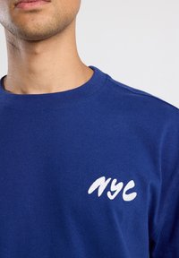 Sininen puuvillaneule, jossa on pyöreä kaula-aukko ja valkoinen "NYC" brodeerattu logo vasemmalla puolella rintaa.