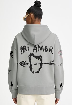 Donna che indossa una felpa grigia con cappuccio con design nero di cuore e freccia e testo "Mi Amor", più grafiche di teschio, fulmine e cuore sulle maniche.