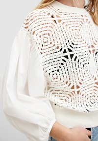 Suéter blanco con un diseño de punto abierto texturizado y patrones geométricos, mangas anchas y una cintura acanalada ajustada.