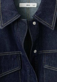 Camicia di jeans blu scuro con colletto a punta, bottoni a pressione argentati e cuciture gialle a contrasto. Due tasche frontali con patta.