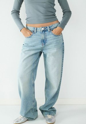 Osoba nosząca luźne, jasnoniebieskie jeansy, srebrne sneakersy i szary długi crop top z rękami w kieszeniach na jednolitym tle.