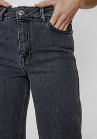 Černé džíny z denimu s vysokým pasem, předními kapsami a kontrastním prošitím. Materiál má hladký, mírně texturovaný vzhled.