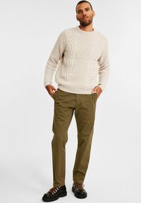 Gebreide trui in lichtbeige met textuurpatronen, gecombineerd met olijfgroene broeken en zwarte veterschoenen met oranje accenten.