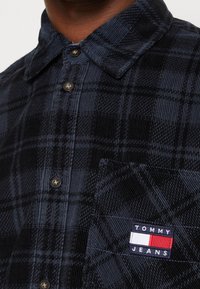 Primo piano di una camicia in cotone a coste blu scuro a scacchi con bottoni neri e una tasca caratterizzata da un etichetta con il logo Tommy Jeans.