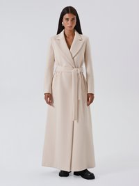 Cappotto lungo beige in tessuto morbido, con design doppiopetto, cintura e tasche laterali. Indossato sopra scarpe platform nere.