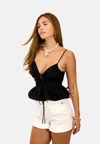 Top peplum negro con cierre de lazo ajustable, que presenta tirantes finos y un escote en V profundo. Combinado con pantalones cortos blancos con dobladillo deshilachado.