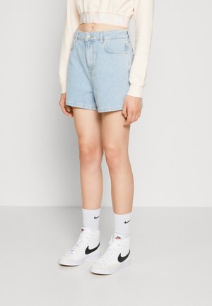 Person trägt hellblaue Jeansshorts, weißes, kurzes Langarmshirt, weiße Nike-Socken und weiße Nike High-Top-Sneaker.