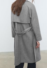 Grijze, wollen blazerjas met een ceintuur in de taille, middellange lengte en brede kraag. Heeft een gelaagd achterontwerp en lange mouwen. Glad textuur.