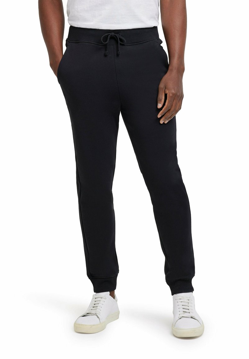 Zwarte trainingsbroek met een trekkoord in de taille, enkelmanchetten en zijzakken, gemaakt van zachte stof. Gepaard met witte sneakers.