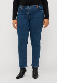 Blue denim jeans with a straight cut, featuring brown accents on the waistband, and a slightly tapered leg. Worn with black heeled boots.
