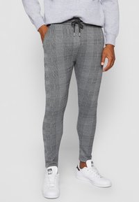 Mężczyzna w szarych, w kratę obcisłych spodniach z czarnym sznurkiem, szarej bluzie i białych trampkach, stojący na jednolitym tle.