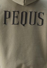 Khaki-Hoodie mit einem großen schwarzen Aufdruck des Wortes "PEQUS" auf dem Rücken; aus weichem, strukturiertem Material gefertigt.
