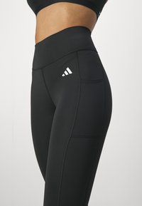 Black high-waisted leggings with a smooth texture, featuring white Adidas logo, side pocket, and structured seams for fit.