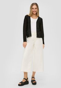 Schwarze, durchsichtige Strickjacke mit strukturierten Streifen, getragen über einem weißen T-Shirt und weiten weißen Hosen. Schwarze Keilsandaletten vervollständigen das Outfit.