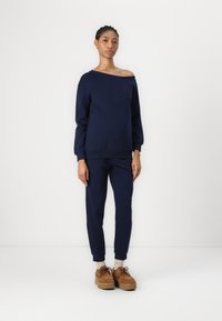 Felpa blu navy con design a spalle scoperte e polsini aderenti, abbinata a pantaloni coordinati. Gli accessori includono scarpe con piattaforma color tan.