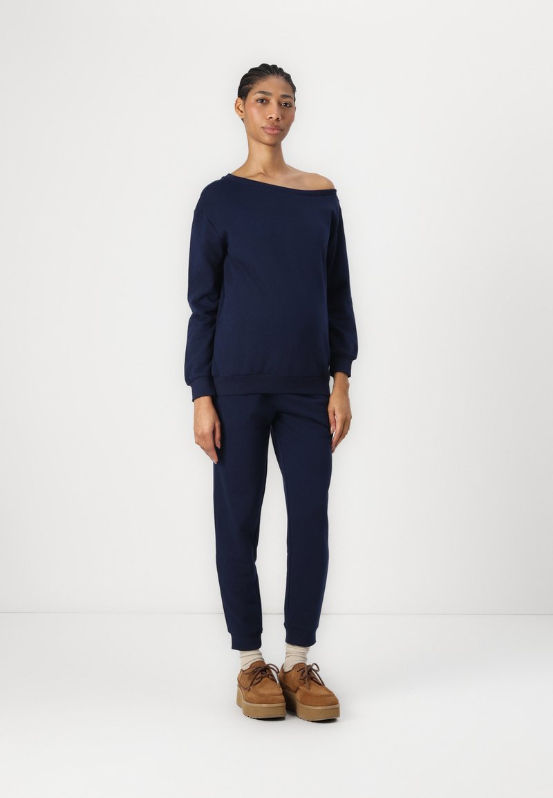Felpa blu navy con design a spalle scoperte e polsini aderenti, abbinata a pantaloni coordinati. Gli accessori includono scarpe con piattaforma color tan.