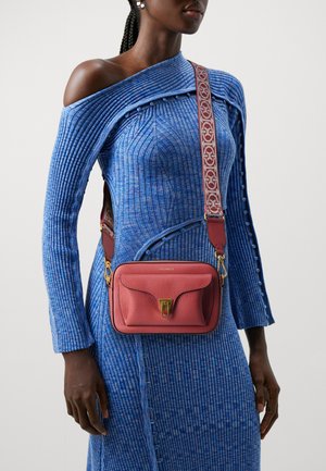 Sac bandoulière en cuir rouge avec un fermoir en or, surface texturée et une sangle à motif. Le modèle porte une robe bleu côtelée à épaules dénudées.