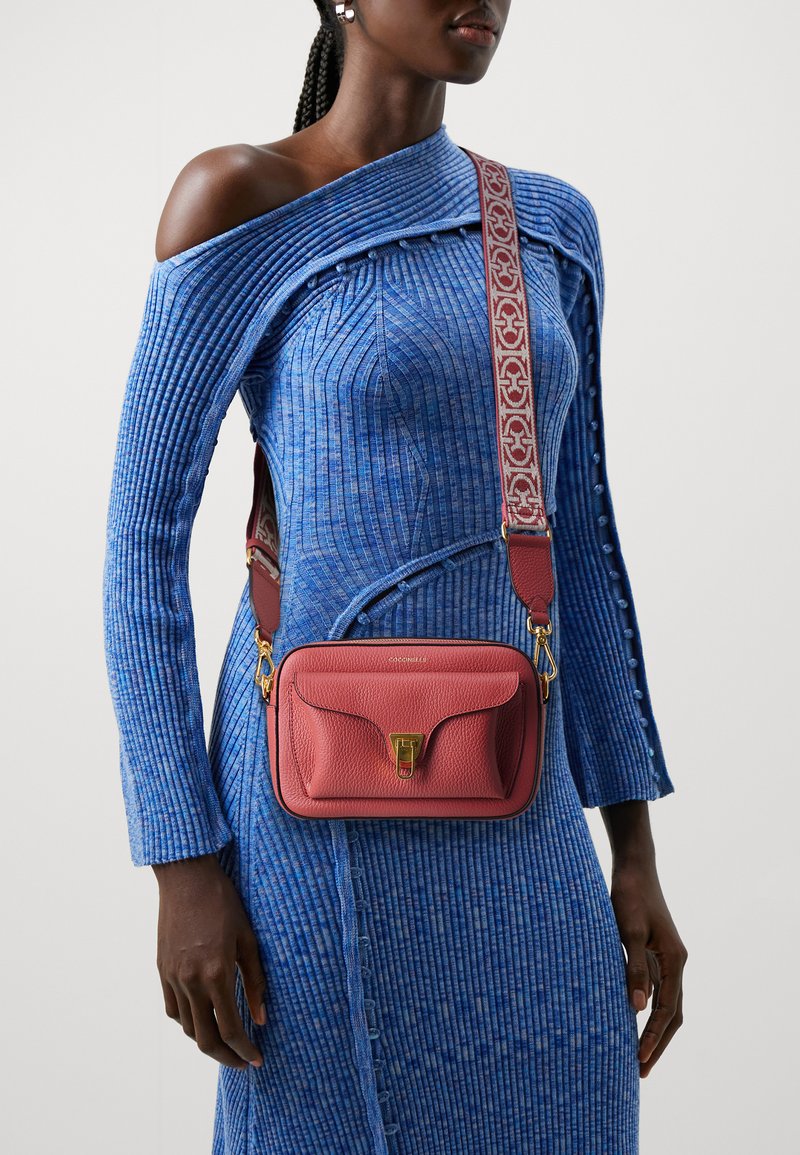 Borsa a tracolla rossa in pelle con chiusura dorata, superficie texture e tracolla con motivo. Il modello indossa un vestito blu a coste con spalle scoperte.
