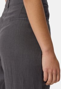 Donkergrijs linnen kledingstuk met een fitted silhouet, voorzien van een zijkeep en een gladde, licht getextureerde oppervlakte. Hand zichtbaar aan de rand.
