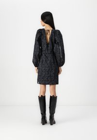Robe noire à motifs avec des manches bouffantes, un dos nu et un détail à nouer, associée à des bottes noires en cuir verni haute jusqu'aux genoux.