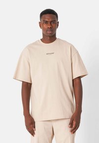 Camiseta de algodón beige con cuello redondo y mangas cortas. Presenta un sutil logo de la marca en una impresión clara en el área del pecho.
