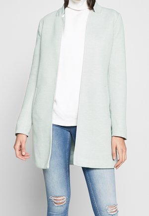 Person trägt einen hellgrauen offenen Mantel, weißen Rollkragenpullover und hellblaue, zerrissene Skinny-Jeans, steht vor neutralem Hintergrund.