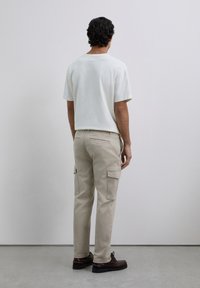 Hombre con pantalones cargo beige, camisa blanca de manga corta y zapatos marrones de pie frente a una pared gris claro lisa.