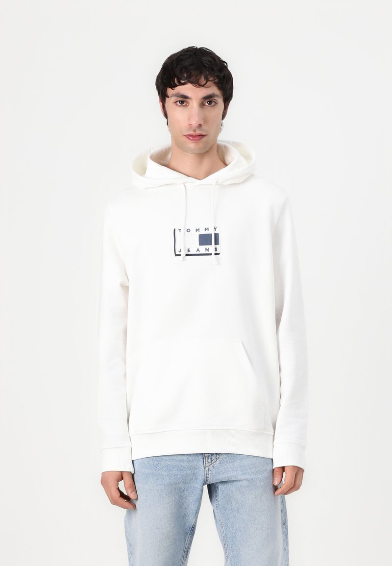 Sweatshirt à capuche blanche en coton, dotée d'une poche kangourou à l'avant et d'un logo graphique bleu marine sur la poitrine. Le jean est de couleur bleu clair.