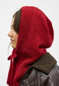 Rød strikket hætte-scarf lavet af blødt uld, med en struktureret overflade og justerbar snøre. Båret over en brun quilted jakke.