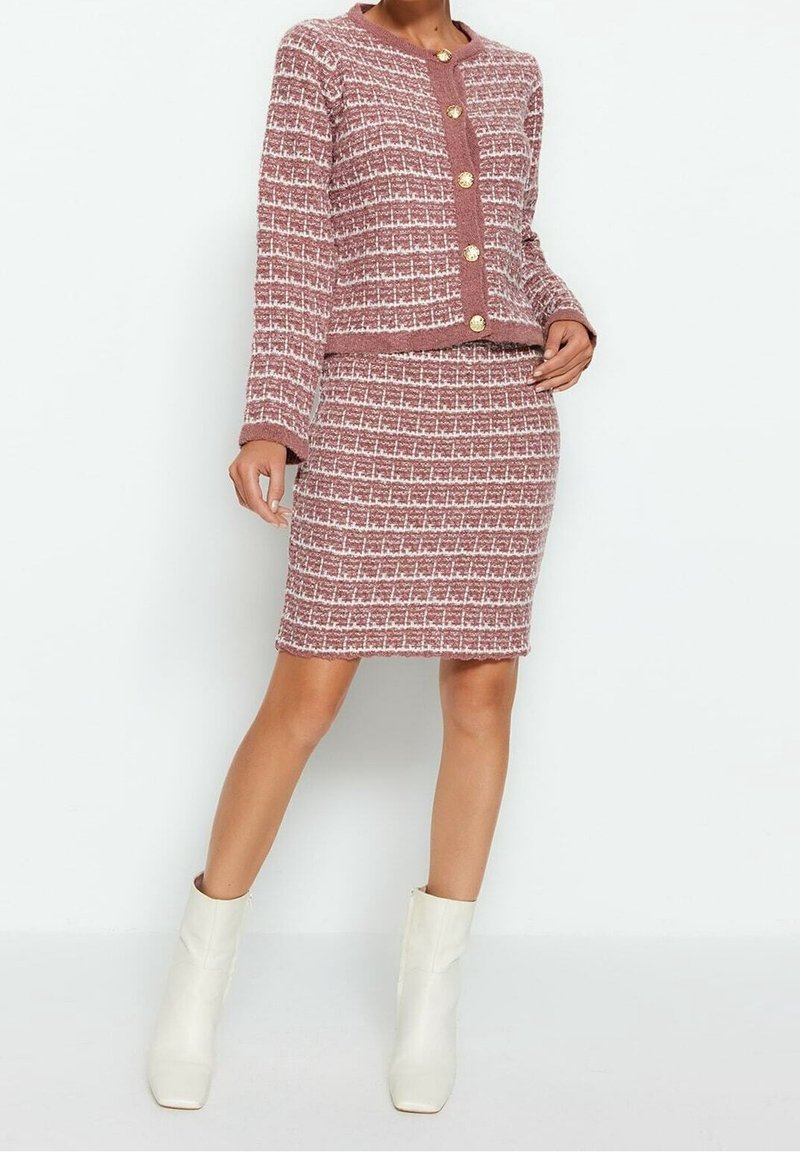 Femme portant une veste texturée rose et blanche à boutons avec une jupe assortie à hauteur du genou et des bottines blanches, debout devant un fond uni.