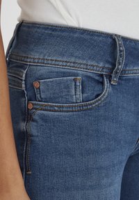 Pantalones vaqueros de mezclilla azul con una cintura de altura media, que presentan costuras en contraste, un pequeño bolsillo delantero y una textura suave.