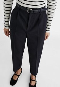 Pantalon noir taille haute sur mesure avec plis à l'avant et ceinture à boucle argentée, associé à un haut rayé à manches longues et des ballerines noires.