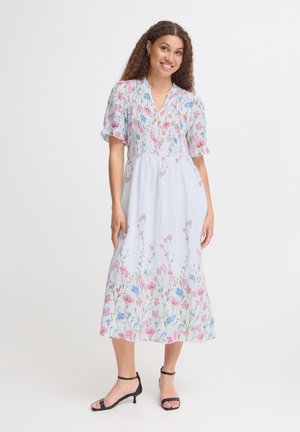 Kvinde med krøllet hår iført hvid mellemlang kjole med pink og blå blomsterprint og sorte ankelrem-sandaler, smilende og stående på hvid baggrund.