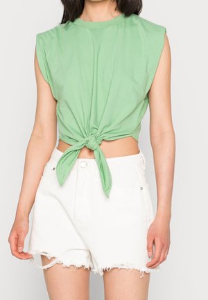 Top senza maniche in cotone verde con design anteriore annodato, abbinato a pantaloni corti bianchi a vita alta con orlo sfrangiato.