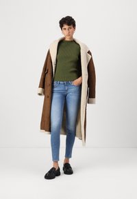 Bruine suède overjas met beige schapenvacht kraag, groene geribbelde trui, blauwe skinny jeans en zwarte loafers. Soepele texturen en neutrale kleuren.