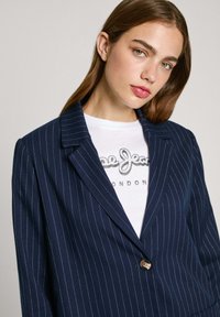 Blazer a rayas azul marino de tela suave, con cierre de botón sencillo y solapas en pico, superpuesto sobre una camiseta blanca.