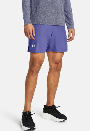 Homme portant un short violet Under Armour, des chaussures de course noires avec semelles blanches et un haut à manches longues bleu, debout sur un fond blanc.