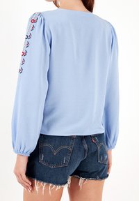 Blouse bleu clair à manches longues bouffantes, ornée d'accents brodés colorés sur les épaules ; portée avec un short en jean foncé.