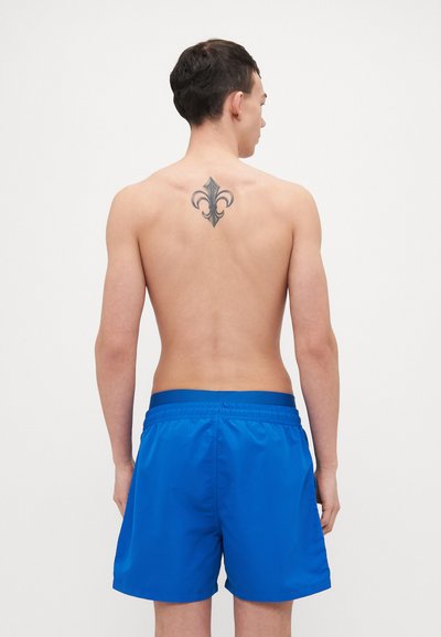 Shorts de bain bleus avec une taille élastique, texture lisse, longueur mi-cuisse et couleur unie. Un tatouage est visible sur le dos du modèle.