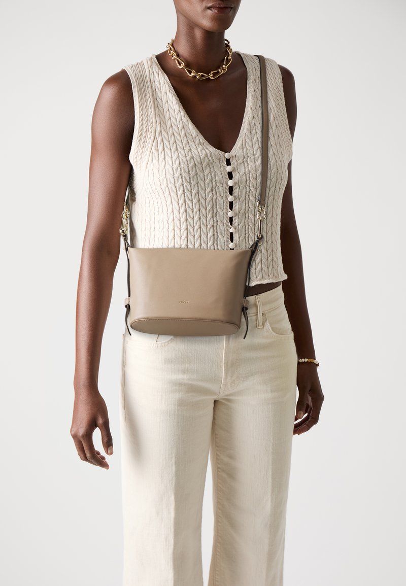 Liten, glatt beige lær crossbody-veske med én stropp, som har subtil merkevare og en ren design. Båret over en lys strikket topp.